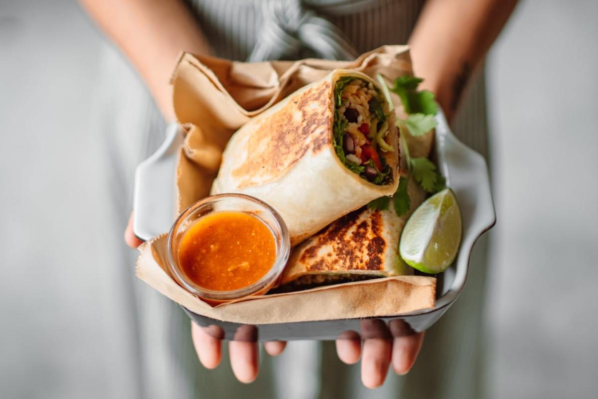 person holding vegetable tortilla wrap with dipping sauce on the side